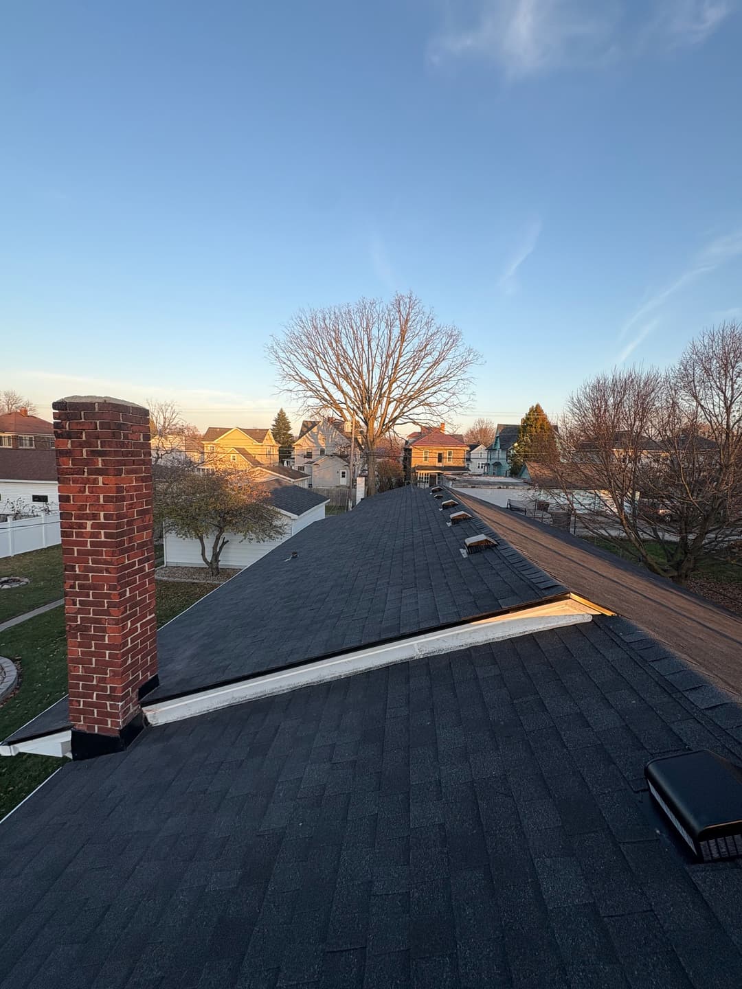 View of a residential roof with chimney, trees, and houses under a clear blue sky.
